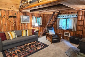 A cabin interior with a wooden interior, a sofa, a chair, a ladder, and a window with curtains.