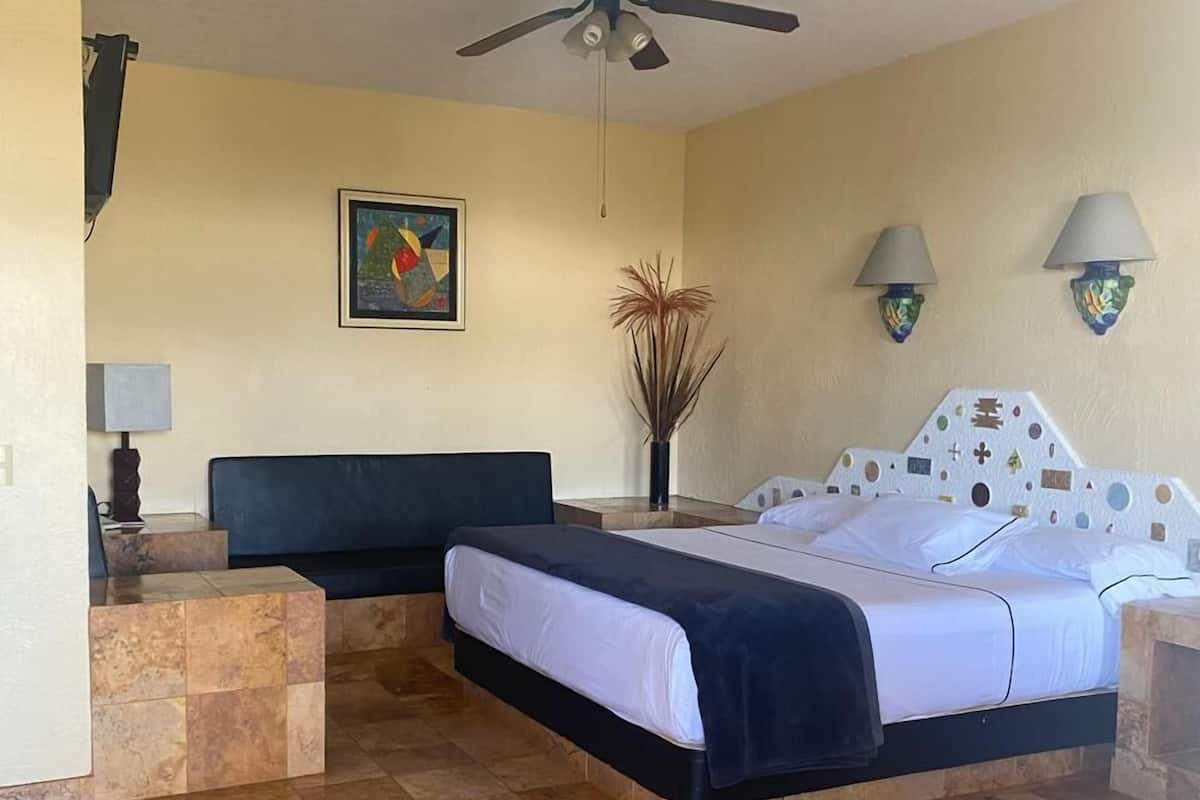 A bedroom with a bed, a ceiling fan, a painting, and a vase with plants.