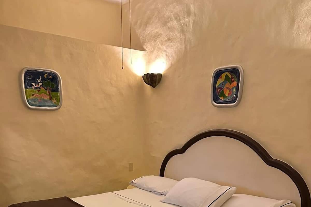 A bed with a brown headboard, white bedding, and a brown throw blanket. There are two wall-mounted light fixtures and two small wall art pieces.