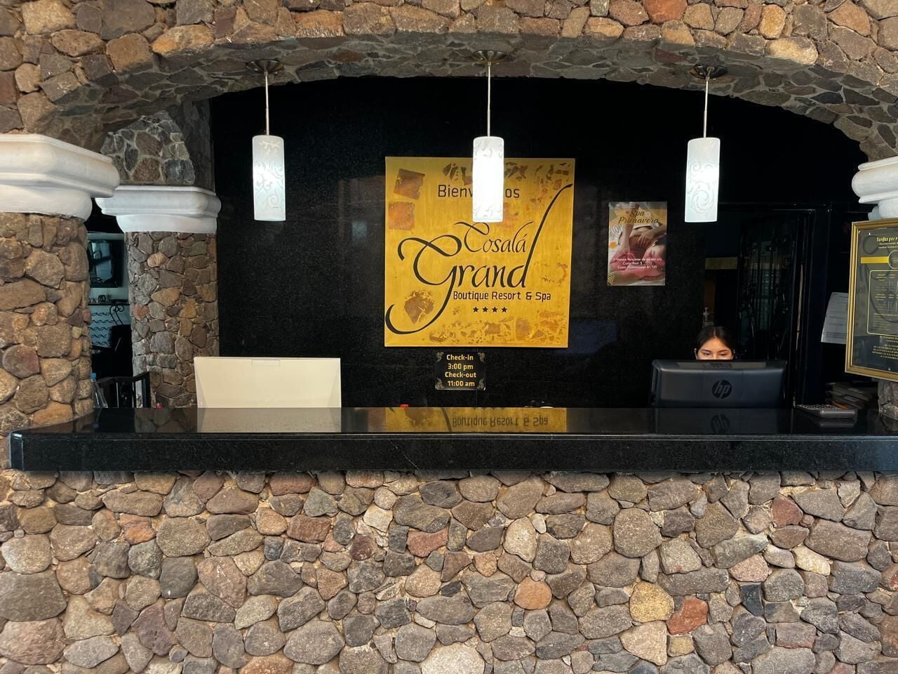 A reception desk with a stone wall backdrop, a sign for 'Cosala Grand Boutique Resort & Spa', and a computer monitor.