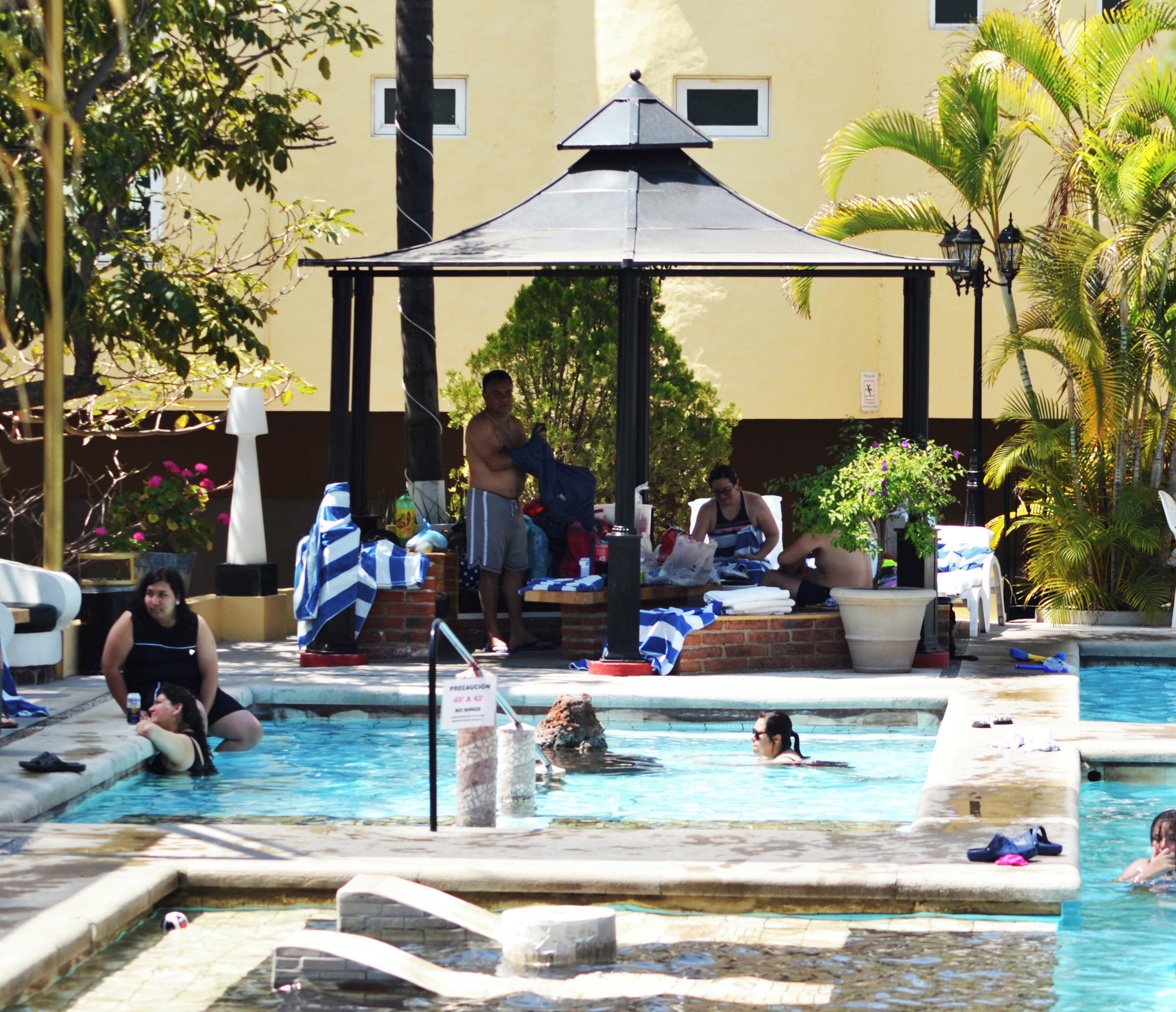 A swimming pool area with lounge chairs and umbrellas, surrounded by palm trees and a building with a tiled roof.