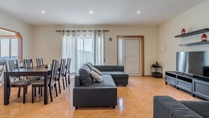 A modern living room with a dining area, grey sofas, a TV, and a large window with curtains. - Casal do Porto - A vacation retreat (Lourido)