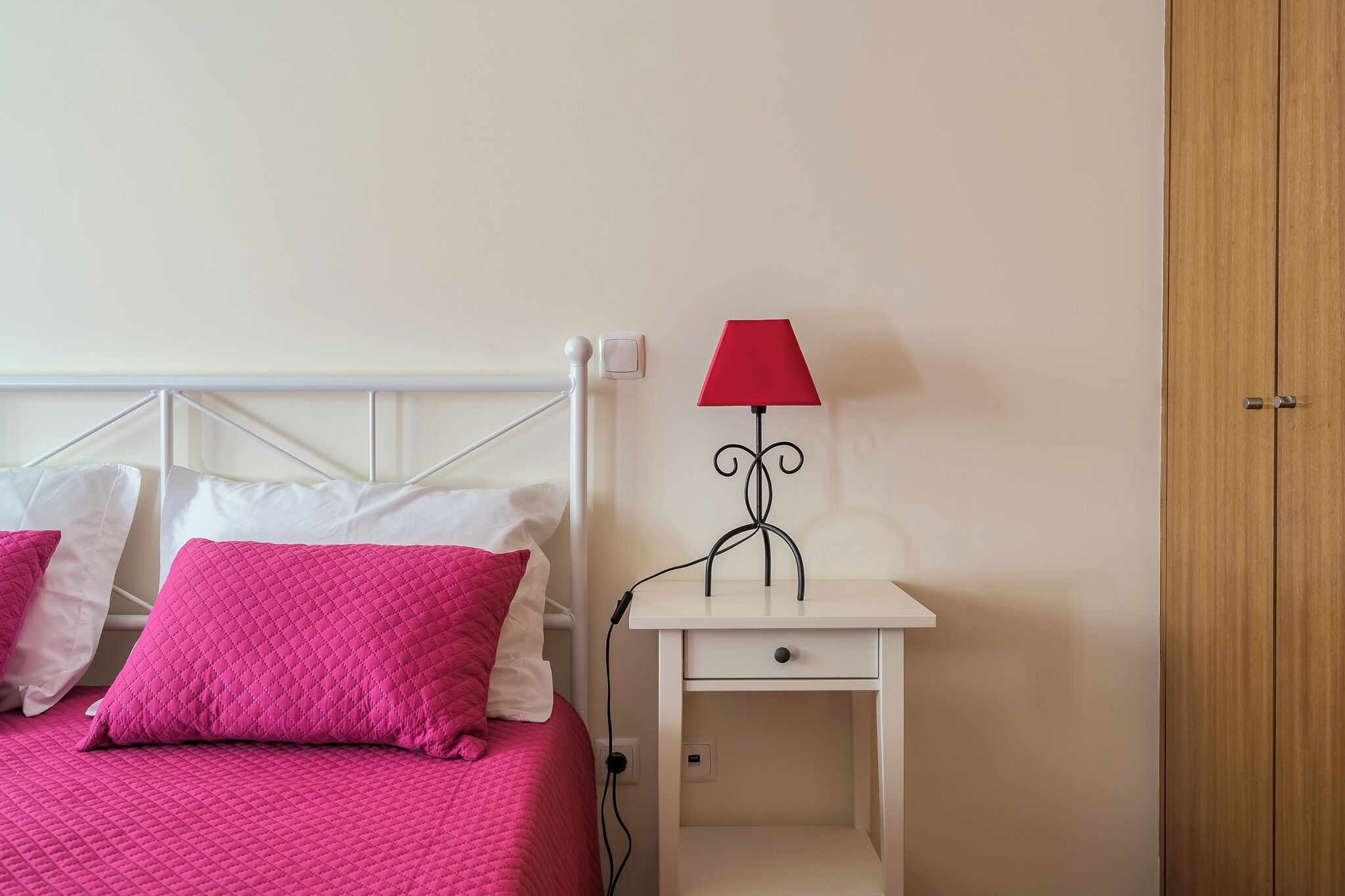 Una cama con ropa de cama rosa, una mesita de noche blanca con una lámpara roja y un armario de madera.