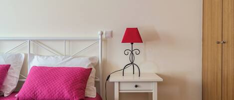 Una cama con ropa de cama rosa, una mesita de noche blanca con una lámpara roja y un armario de madera.