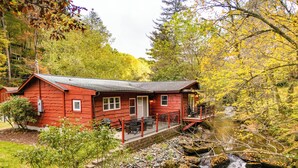 A red cabin with a porch overlooking a stream, surrounded by autumn foliage.