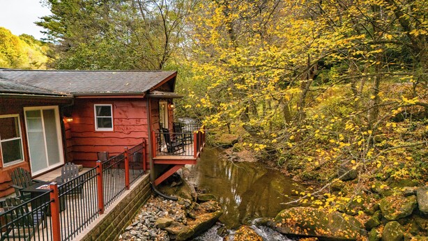 A red cabin with a deck overlooking a stream, surrounded by autumn foliage.