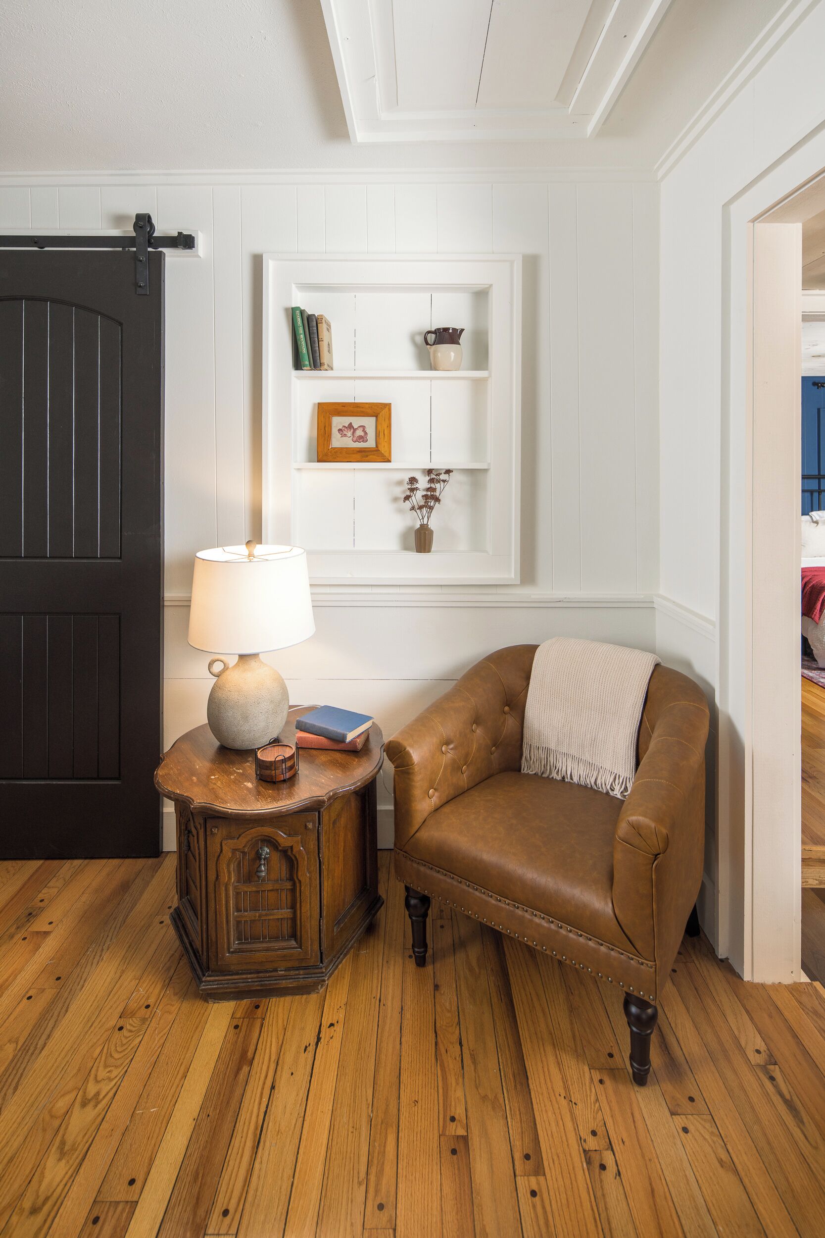 A room with a wooden floor, a dark door, a lamp on a small wooden table, a leather armchair, and a white shelf with books and a vase.