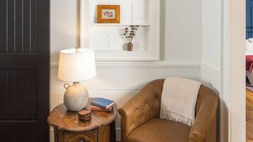 A room with a wooden floor, a dark door, a lamp on a small wooden table, a leather armchair, and a white shelf with books and a vase.