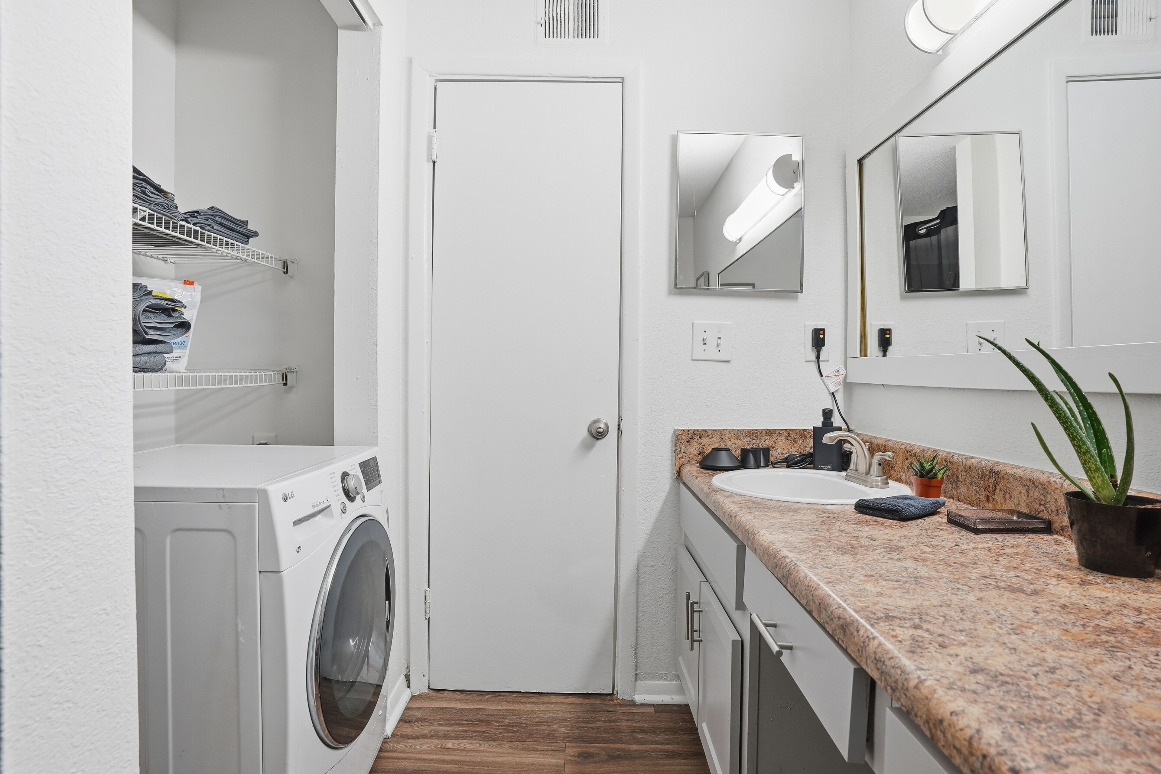 A compact laundry room with a washing machine, shelves with folded clothes, a sink, and a mirror.