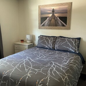 Un dormitorio con cama, mesita de noche, lámpara, un cuadro en la pared y vista a un faro.
