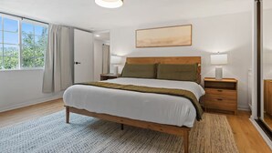 A bedroom with a wooden bed, a bedside table with a lamp, a window with curtains, and a painting on the wall.