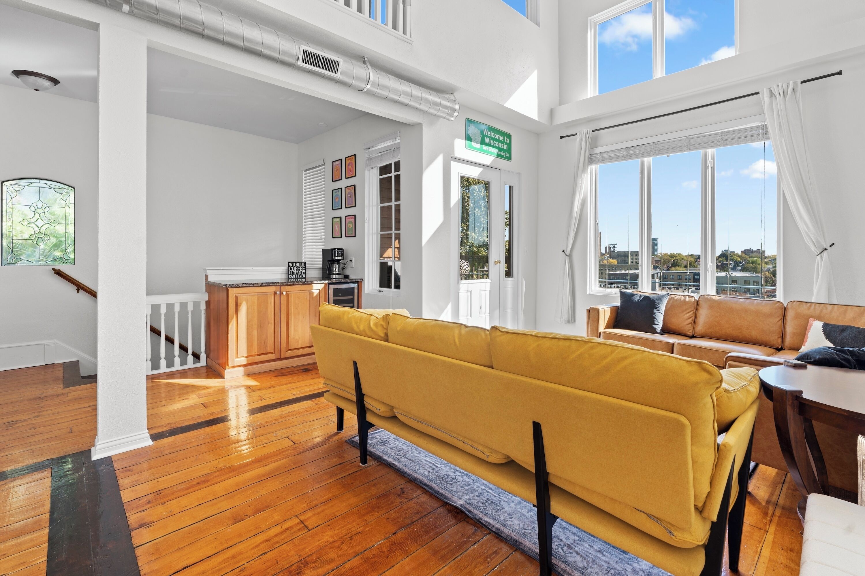 Uma sala de estar ampla com sofá amarelo, piso de madeira, escada e vista para a cidade.
