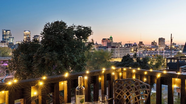 A cityscape view from a balcony with string lights, a bottle of liquor, and a glass on a table.