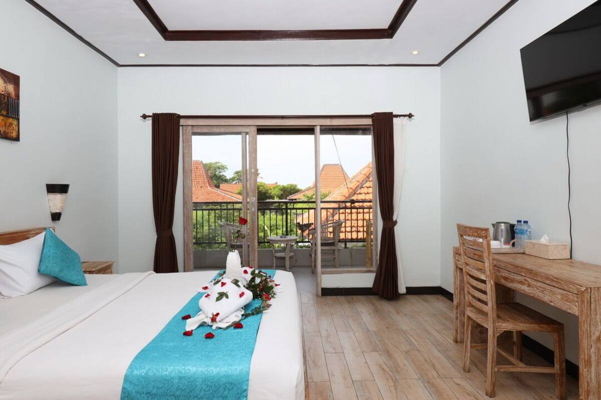 A hotel room with a bed, a desk, a chair, a balcony with a view of houses, and a television on the wall.