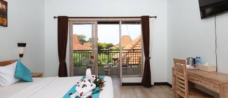 Een hotelkamer met een bed, een bureau, een stoel, een balkon met uitzicht op huizen, en een televisie aan de muur.