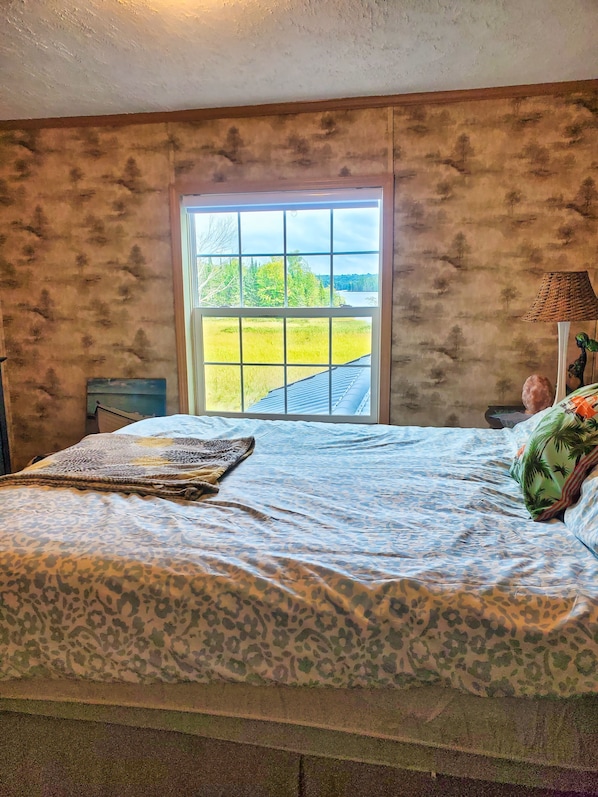 Een slaapkamer met een groot raam, een bed met een luipaardprint deken en uitzicht op een grasveld en bomen.