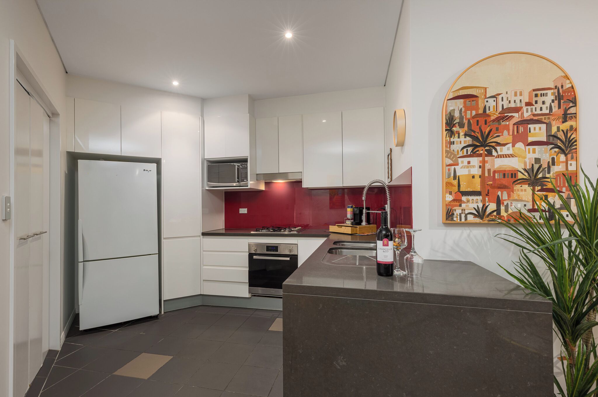 A modern kitchen with white cabinets, a red backsplash, a microwave, a stove, and a wine bottle on the counter.