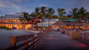 A beachfront hotel with a wooden boardwalk leading to the entrance, palm trees, and thatched-roof structures.