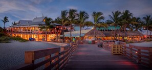 A beachfront hotel with a wooden boardwalk leading to the entrance, palm trees, and thatched-roof structures.