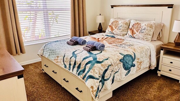 A bedroom with a bed, a ceiling fan, a nightstand, a lamp, and a window with curtains.