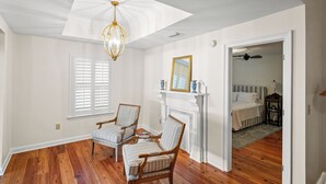 Una habitación amplia con pisos de madera, una chimenea blanca, dos sillones a rayas y un ventilador de techo, además de una cama en la habitación contigua.