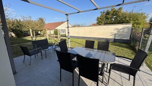 Een terras buiten met een tafel en stoelen, omringd door een metalen hek en uitzicht op een huis en bomen.