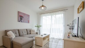 Een moderne woonkamer met een bank, een salontafel, een televisie en een groot raam met dunne gordijnen.