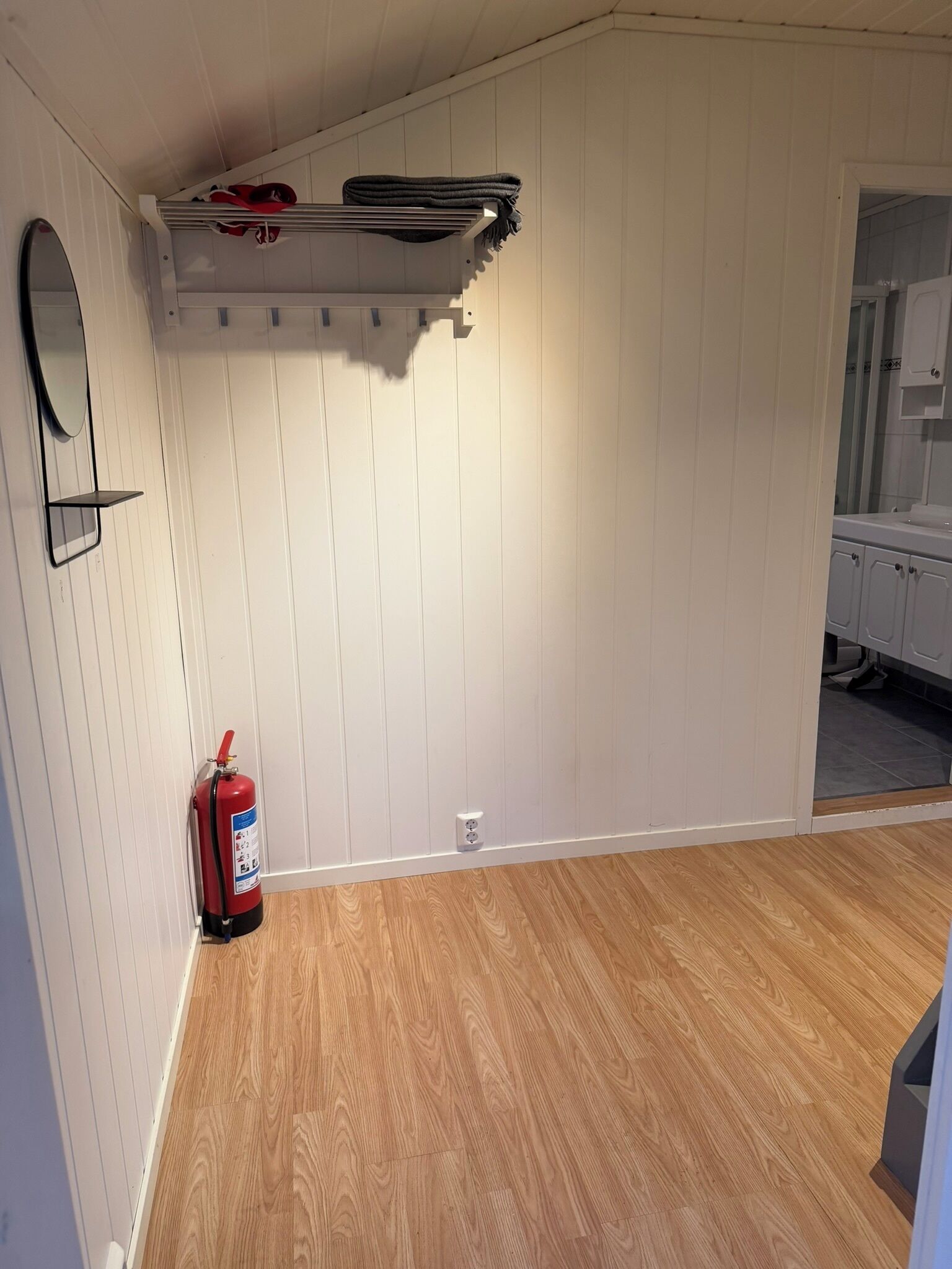 A room with white paneled walls, a wooden floor, a fire extinguisher, and a mirror on the left wall.