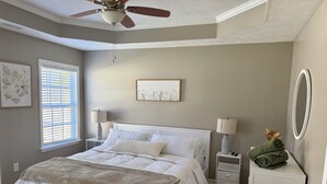 Ein Schlafzimmer mit Bett, Nachttischen, Deckenventilator, Fenster mit Jalousien, einem gerahmten Bild und einem Spiegel.