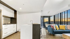 Een moderne keuken met witte kasten en een centraal eiland, een zithoek met een blauwe bank en kleurrijke kussens, en grote ramen met uitzicht op de stad.