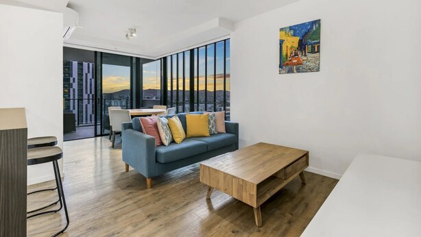 Een moderne woonkamer met een blauwe bank, een houten salontafel en uitzicht op de stad door grote ramen.