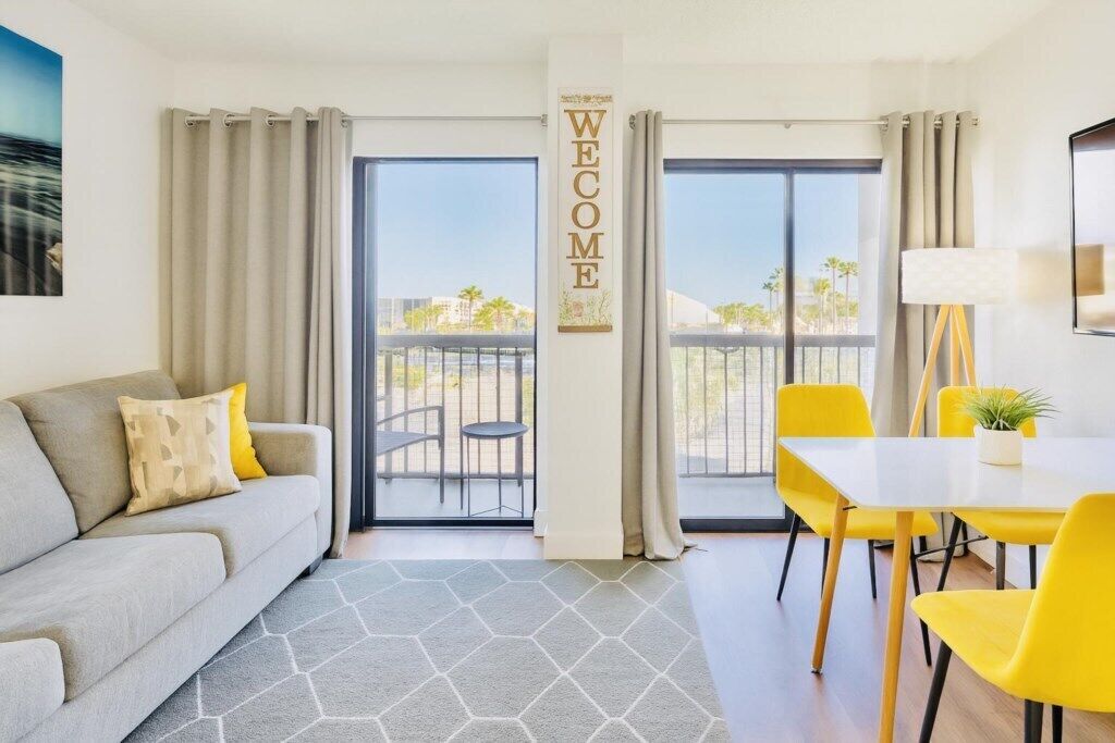Una habitación de hotel moderna con un sofá, una mesa de comedor con sillas amarillas, vistas al exterior a través de amplios ventanales y un cartel que dice 'WELCOME'.