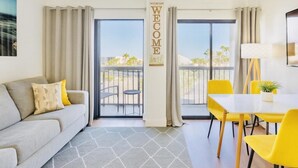 A modern hotel room with a sofa, a dining table with yellow chairs, a view of the outdoors through large windows, and a 'WELCOME' sign. - Spacious 2BR Orlando Suite | Full Kitchen & Balcony Near Epic & Universal (Orlando)