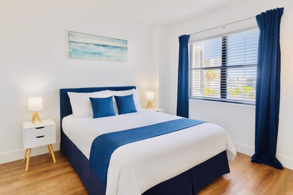 Habitación de hotel con cama, mesita de noche, lámpara, ventana con cortinas y un cuadro en la pared.