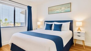 A neatly made bed with a blue and white bedspread, two bedside tables with lamps, a window with curtains, and a painting of sailboats on the wall. - Spacious 2BR Orlando Suite | Full Kitchen & Balcony Near Epic & Universal (Orlando)