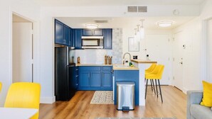 A modern kitchen with blue cabinets, a black refrigerator, and a small island with a sink. There are yellow chairs and a gray rug on the floor. - Spacious 2BR Orlando Suite | Full Kitchen & Balcony Near Epic & Universal (Orlando)