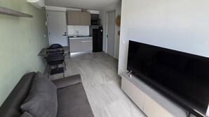 A modern living room with a flat-screen TV, a sofa, and a dining area with a kitchen in the background. - Modern Retreat 1- Bed T3-529 (Chalong)