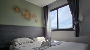 A neatly made bed with towels arranged in a swan shape, a large window with curtains, and wall decor including sunburst patterns. - Modern Retreat 1- Bed T3-529 (Chalong)