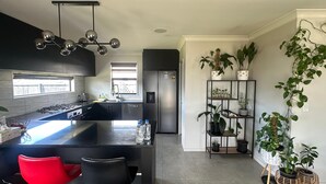 A modern kitchen with a central island, black cabinetry, and a stainless steel refrigerator. There are potted plants on shelves and a red chair.