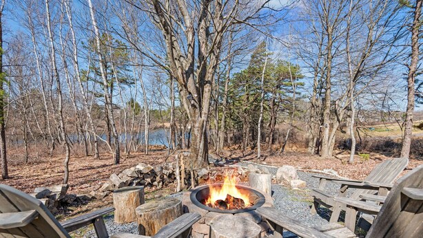 A fire pit surrounded by wooden chairs in a forested area with a lake in the background.