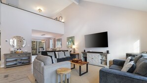 A spacious living room with a grey sofa, a coffee table, and a TV mounted on the wall.