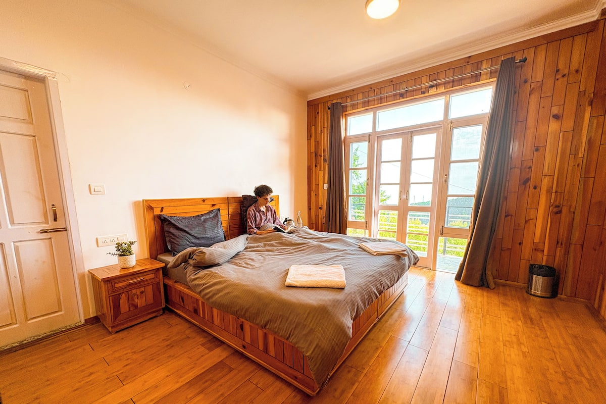 A wooden-framed bed with a headboard, a bedside table with a plant, and a window with curtains.