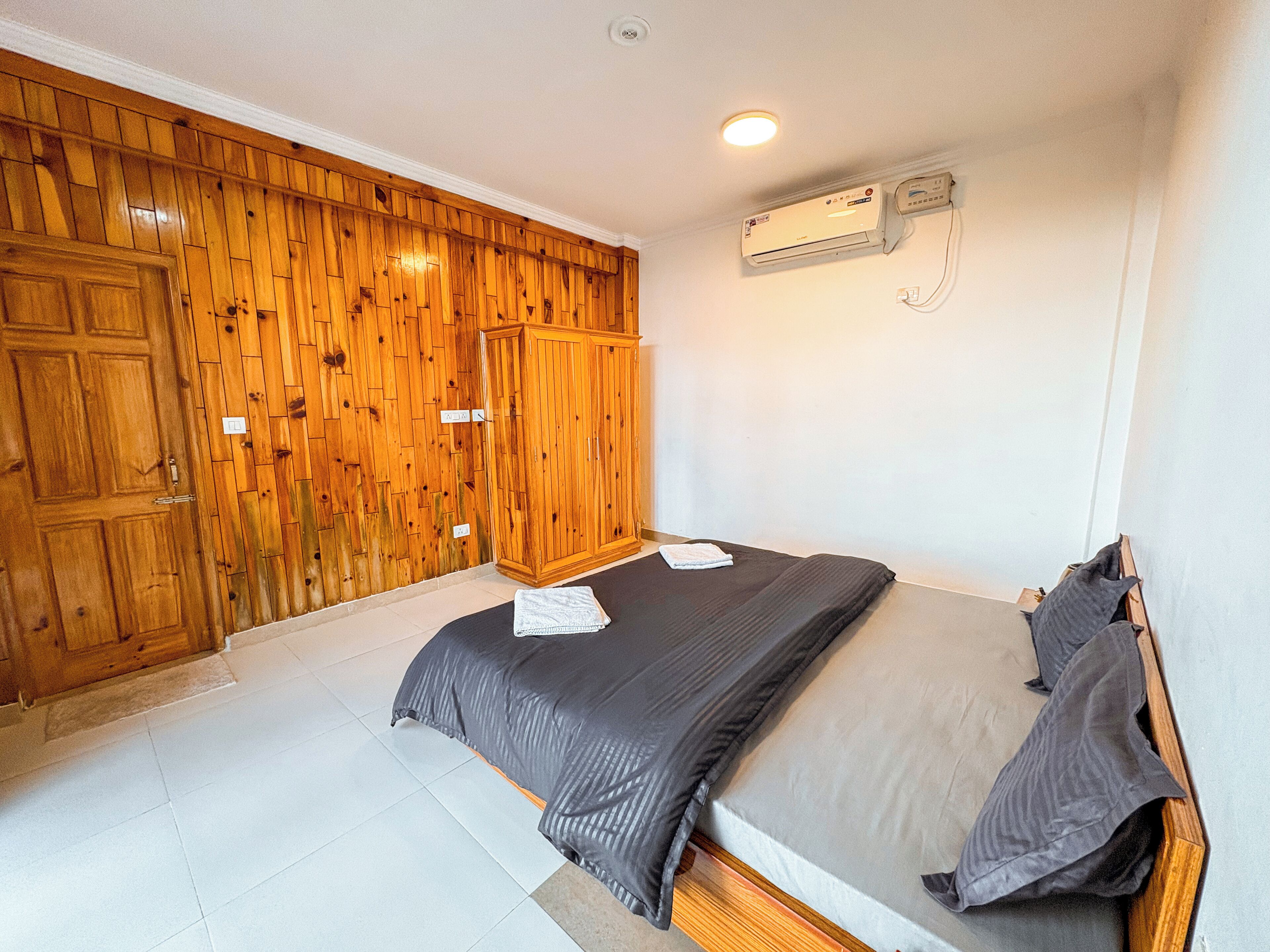 A bedroom with a wooden wardrobe, a bed with grey bedding, and an air conditioning unit on the wall.