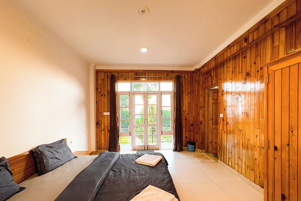 A bedroom with a large bed, wooden paneling on the walls, and a view through a glass door to the outside.