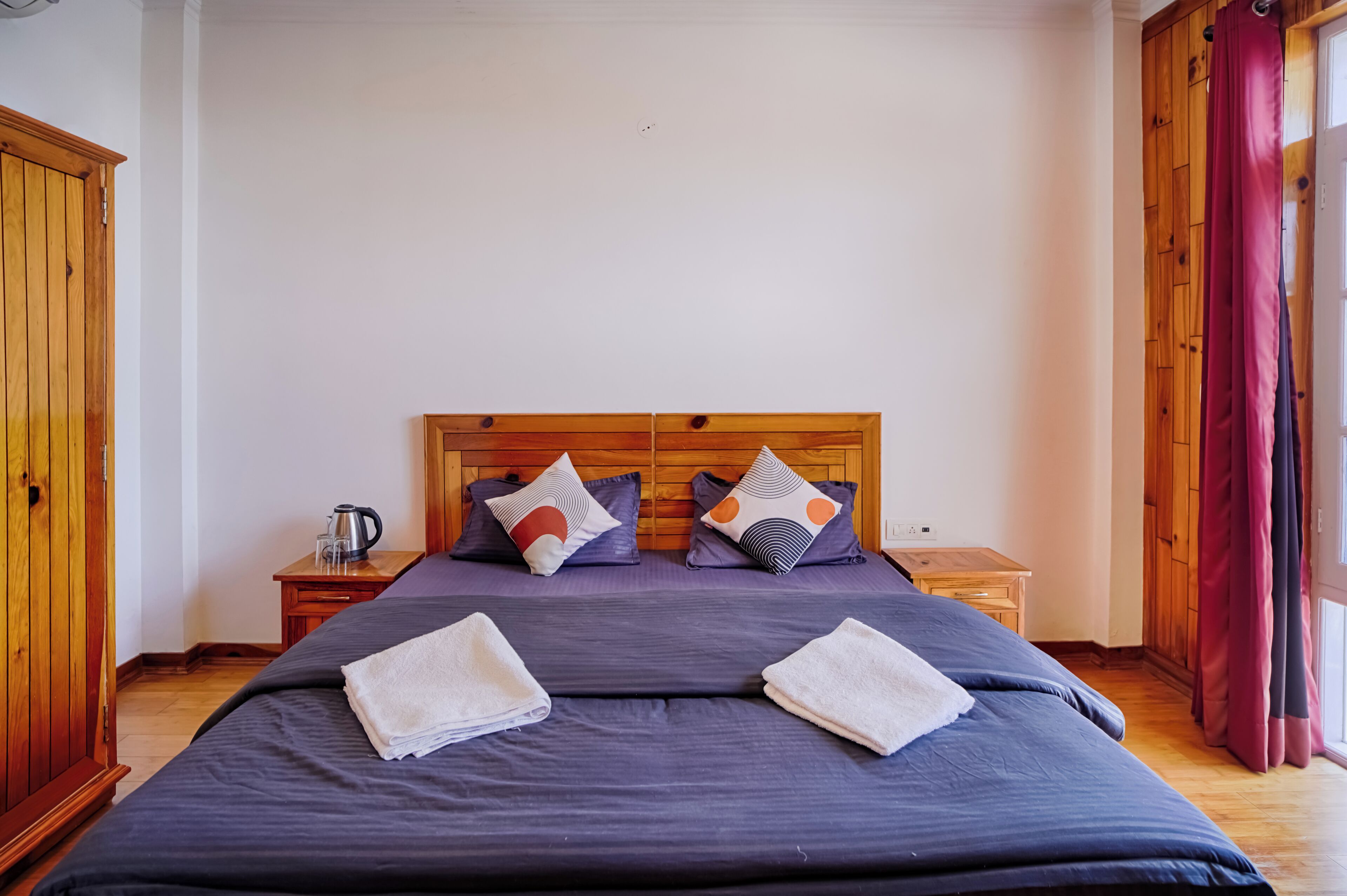 A double bed with a wooden headboard, two nightstands, a kettle, and a window with curtains.