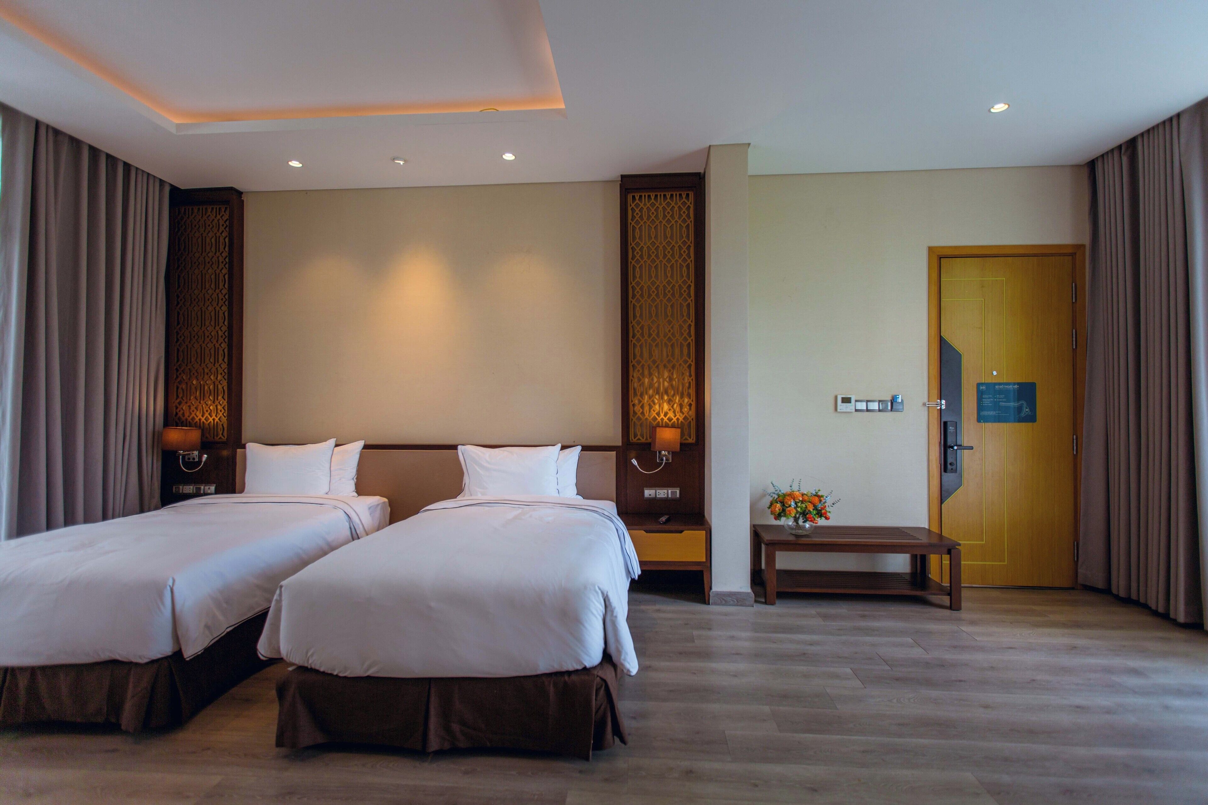 A hotel room with two beds, a wooden floor, a nightstand with a lamp, and a door with a sign.