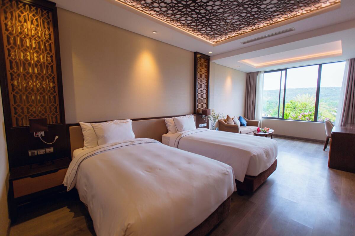A hotel room with two beds, a desk, a chair, and a large window offering a view of greenery.