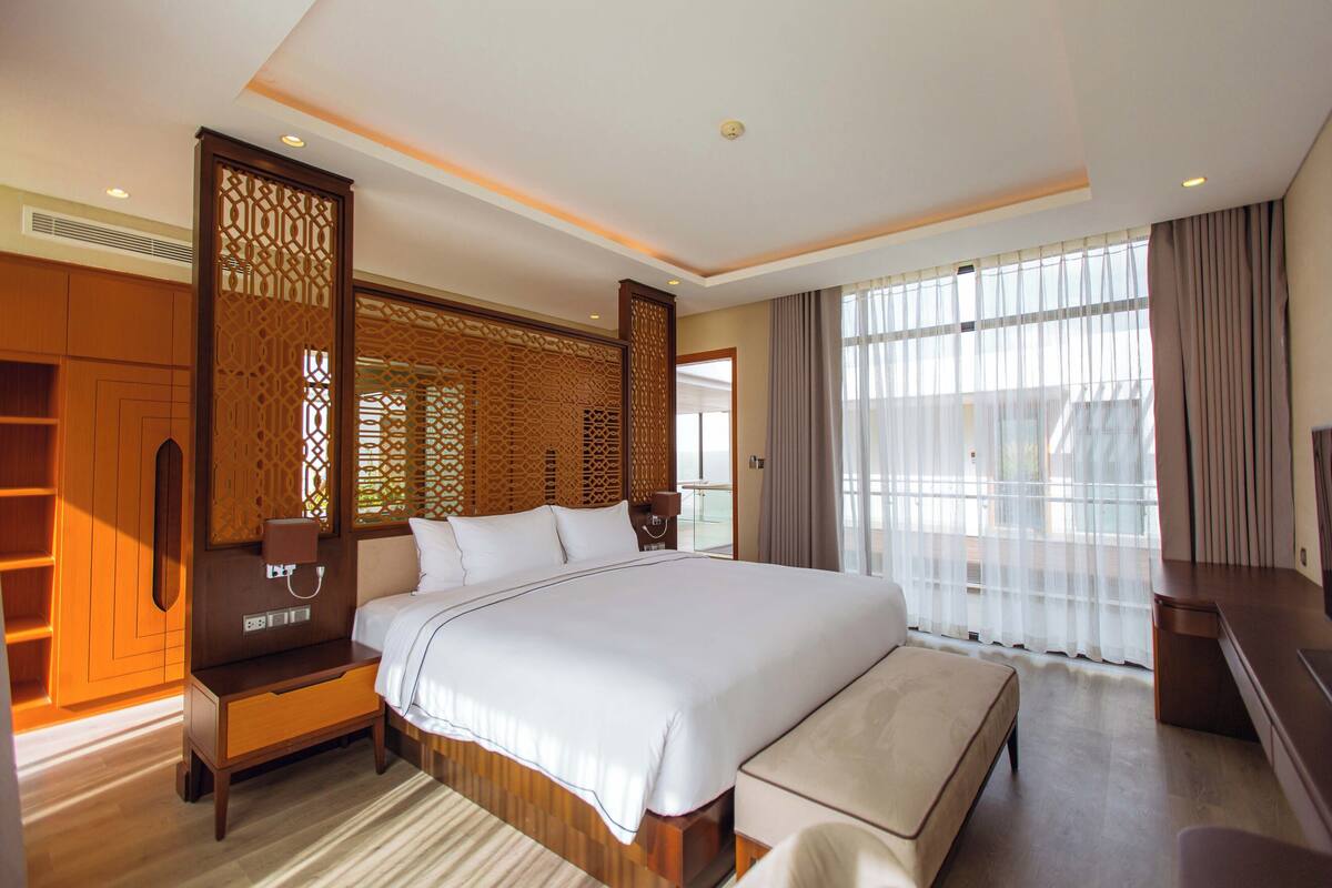 A spacious hotel room with a large bed, a wooden wardrobe, a desk, and a large window with curtains.