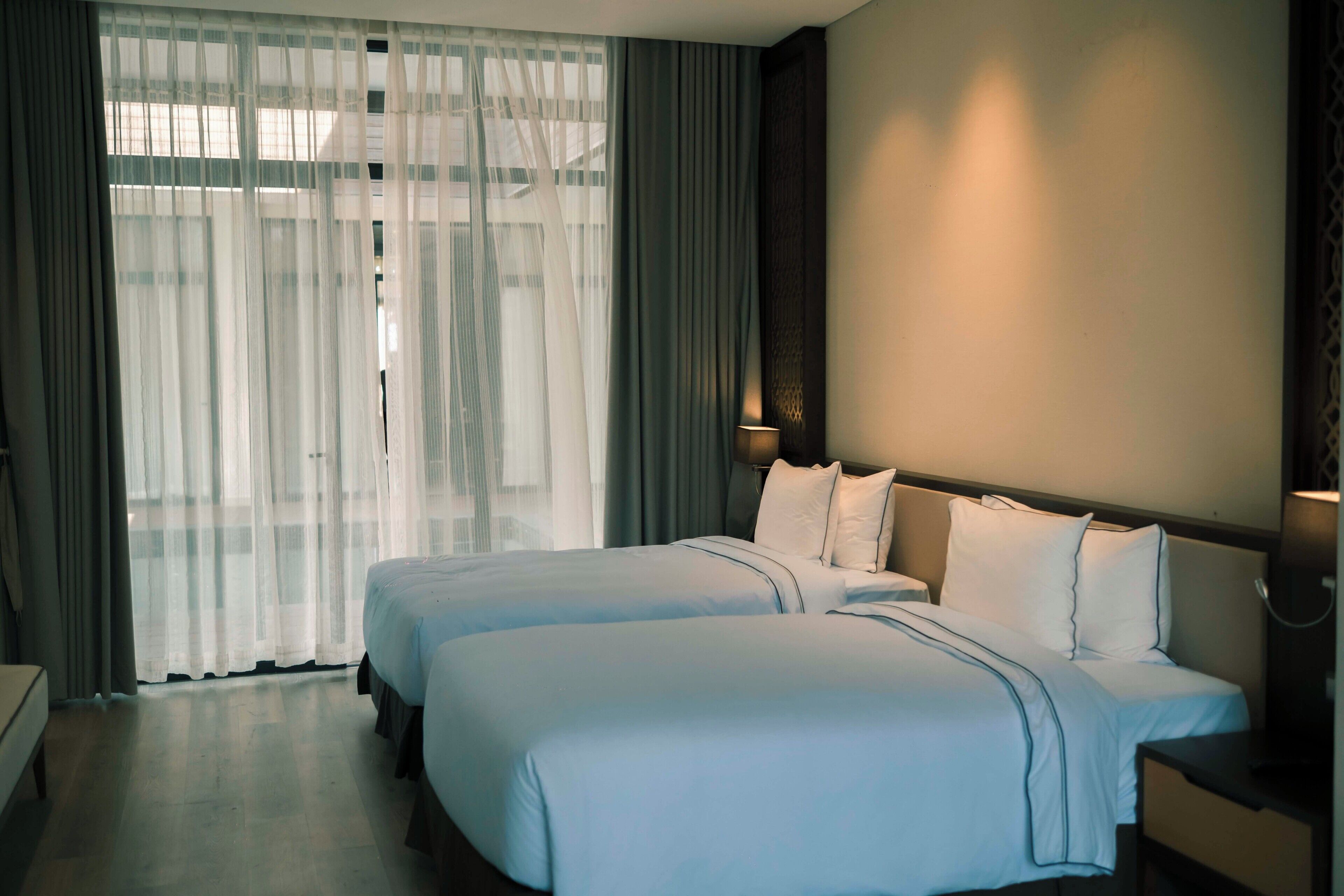 Ein Hotelzimmer mit zwei Betten, einem großen Fenster mit transparenten Vorhängen, einem Nachttisch mit Lampe sowie einer Wandleuchte.
