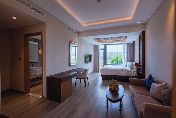 A modern hotel room with a desk, a bed, a sofa, and a view of the outdoors.